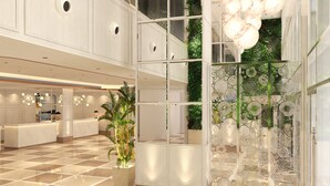 Un moderno vestíbulo de hotel con techo alto, grandes candelabros, una mesa de recepción y un separador de vidrio con plantas.