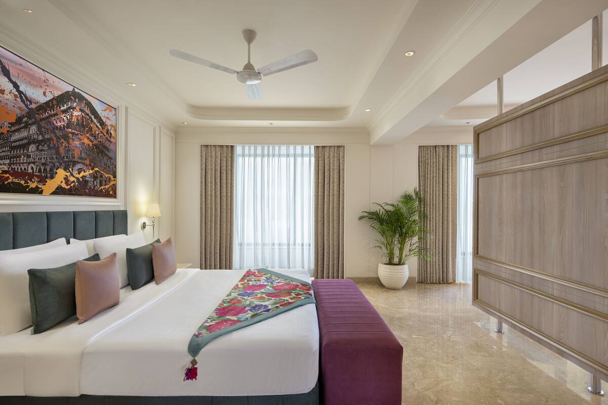 Une chambre d’hôtel avec un grand lit, un ventilateur de plafond, une grande fenêtre avec des rideaux, une plante en pot et un mur recouvert de panneaux de bois.