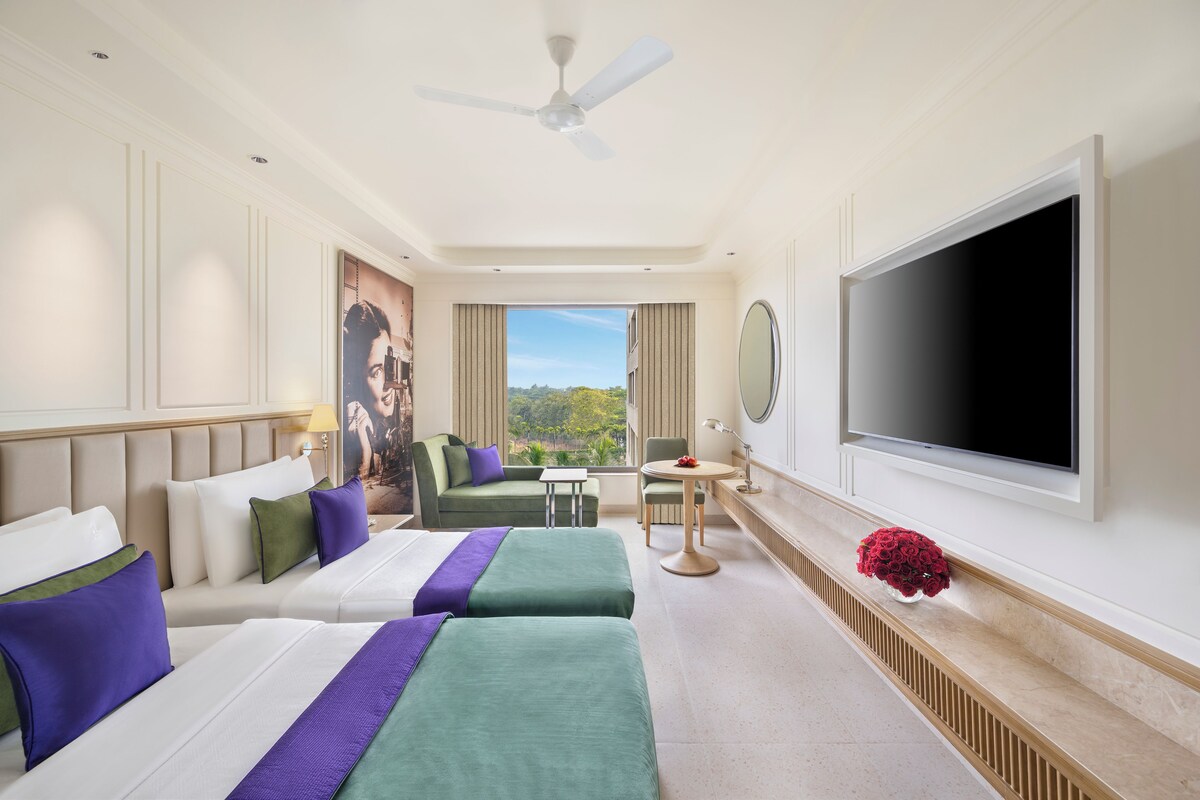 Hotelkamer met twee bedden, een televisie, plafondventilator, raam met uitzicht en een kleintje tafel met een bloemenvaas.