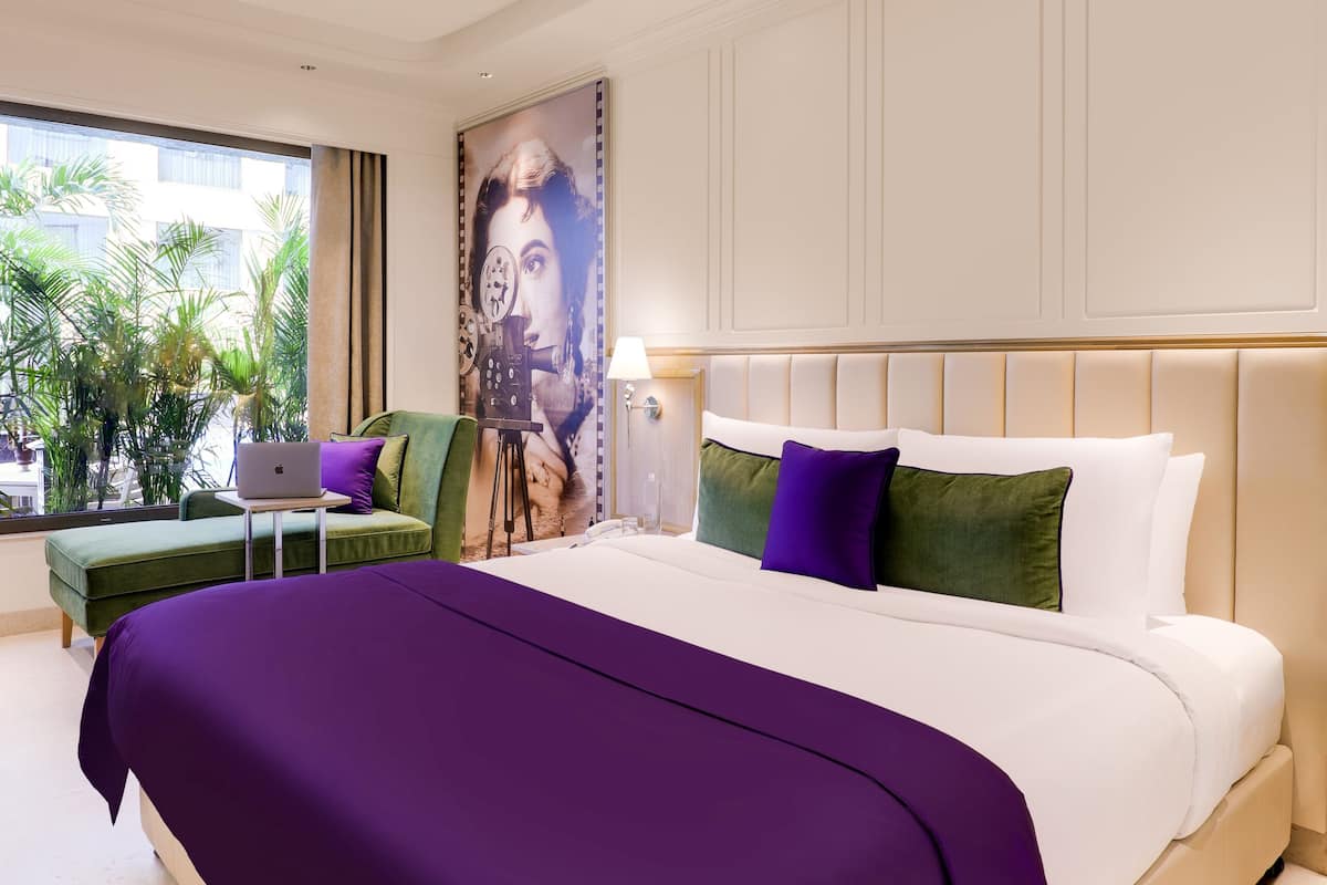Une chambre d’hôtel avec un grand lit, un bureau avec un ordinateur portable, un fauteuil vert et une vue sur les palmiers par la fenêtre.
