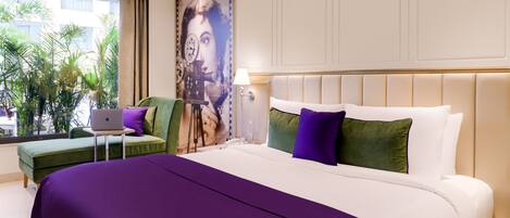 Habitación de hotel con una cama grande, un escritorio con una computadora portátil, un sillón verde y vistas a palmeras a través de la ventana.
