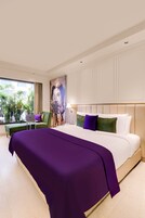 A hotel room with a large bed, a desk with a laptop, a green armchair, and a view of palm trees through the window.
