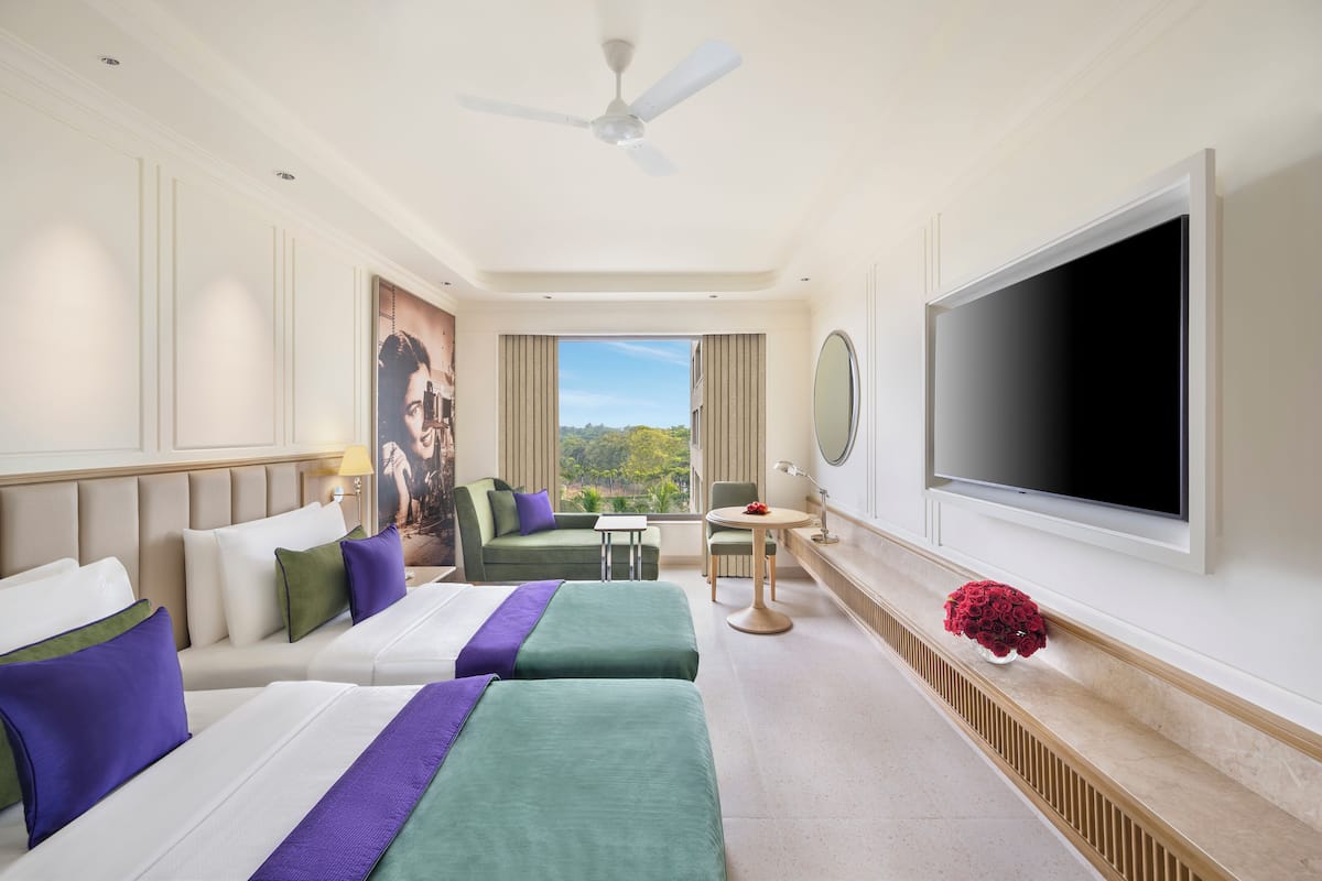 Une chambre d’hôtel avec deux lits, une télévision, un ventilateur de plafond, une fenêtre avec vue et une petite table sur laquelle est posé un vase de fleurs.