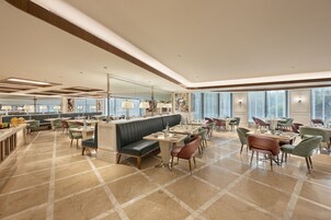 Een ruime hotellobby met een centraal zitgedeelte, diverse eettafels en grote ramen die natuurlijk licht binnenlaten.