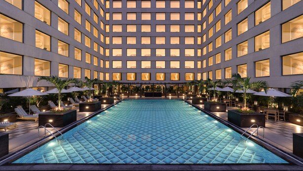 Zona de piscina con sillas de descanso y sombrillas, rodeada por un edificio con múltiples ventanales.