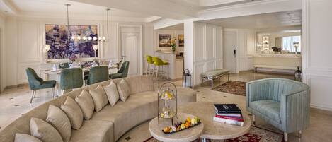 A spacious living room with a large beige sectional sofa, a round coffee table with snacks, and a dining area with green chairs and a chandelier.