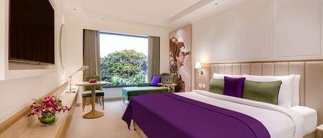 A modern hotel room with a large bed, a desk, and a view of greenery outside.