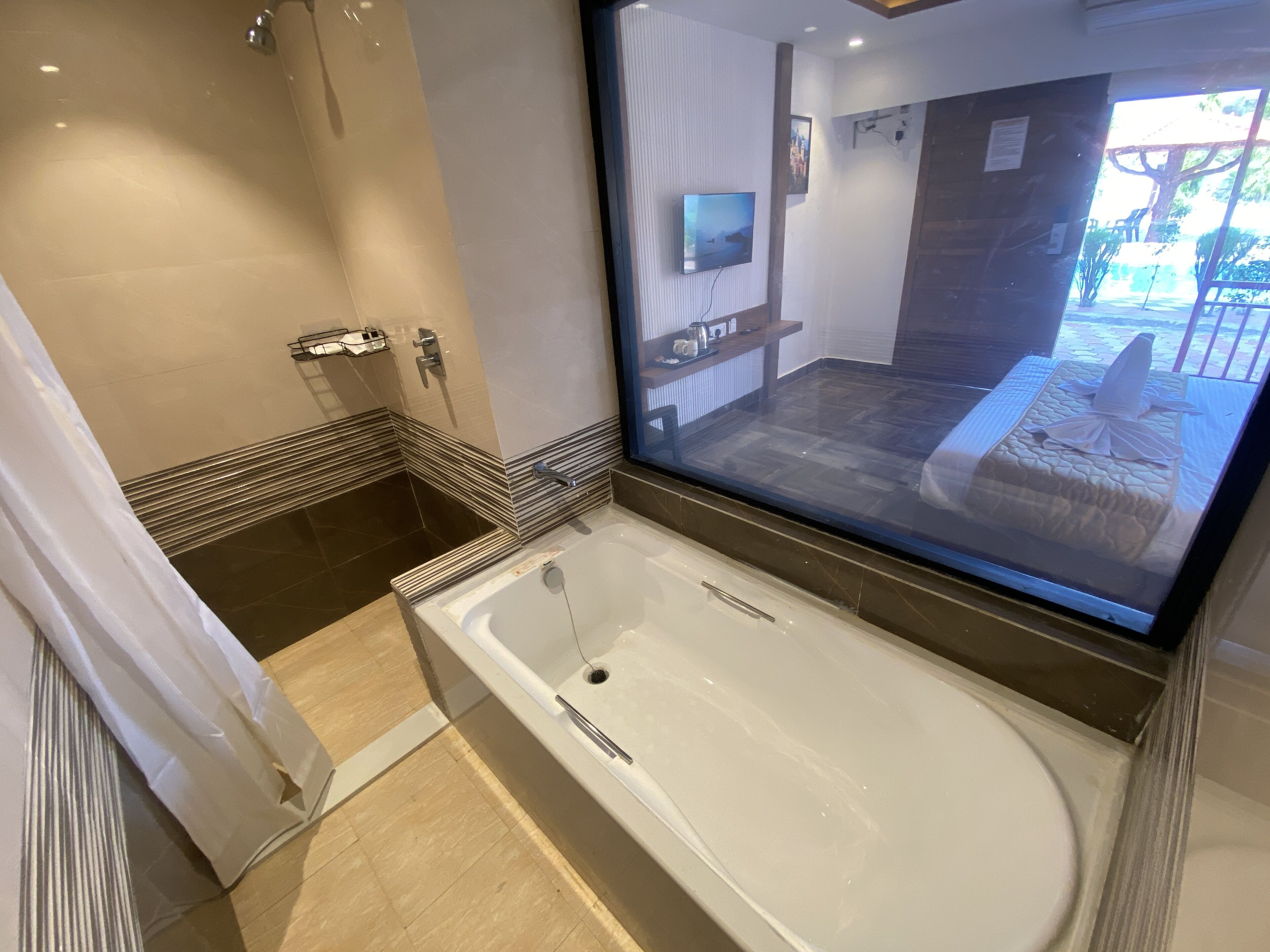 A modern bathroom with a large bathtub, a shower area, and a mirror reflecting a bedroom with a bed and a TV.