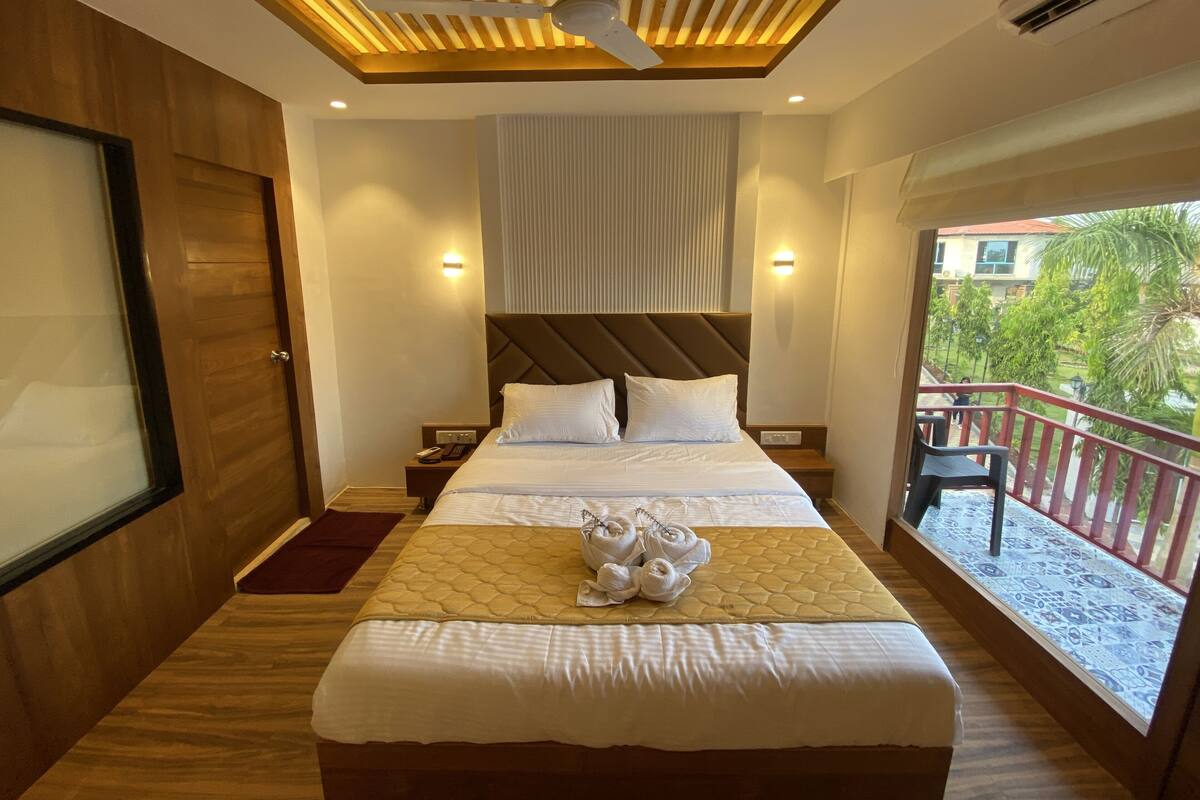 A hotel room with a bed, a ceiling fan, a nightstand, a window with a view of greenery, and a balcony with a railing.