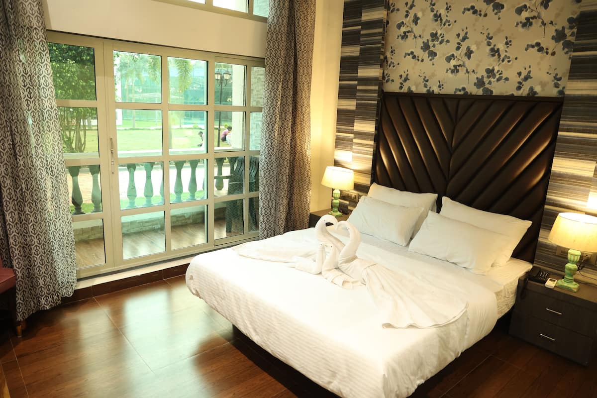 A hotel room with a large window, a bed with white linens, a headboard with a floral pattern, and a nightstand with a lamp.