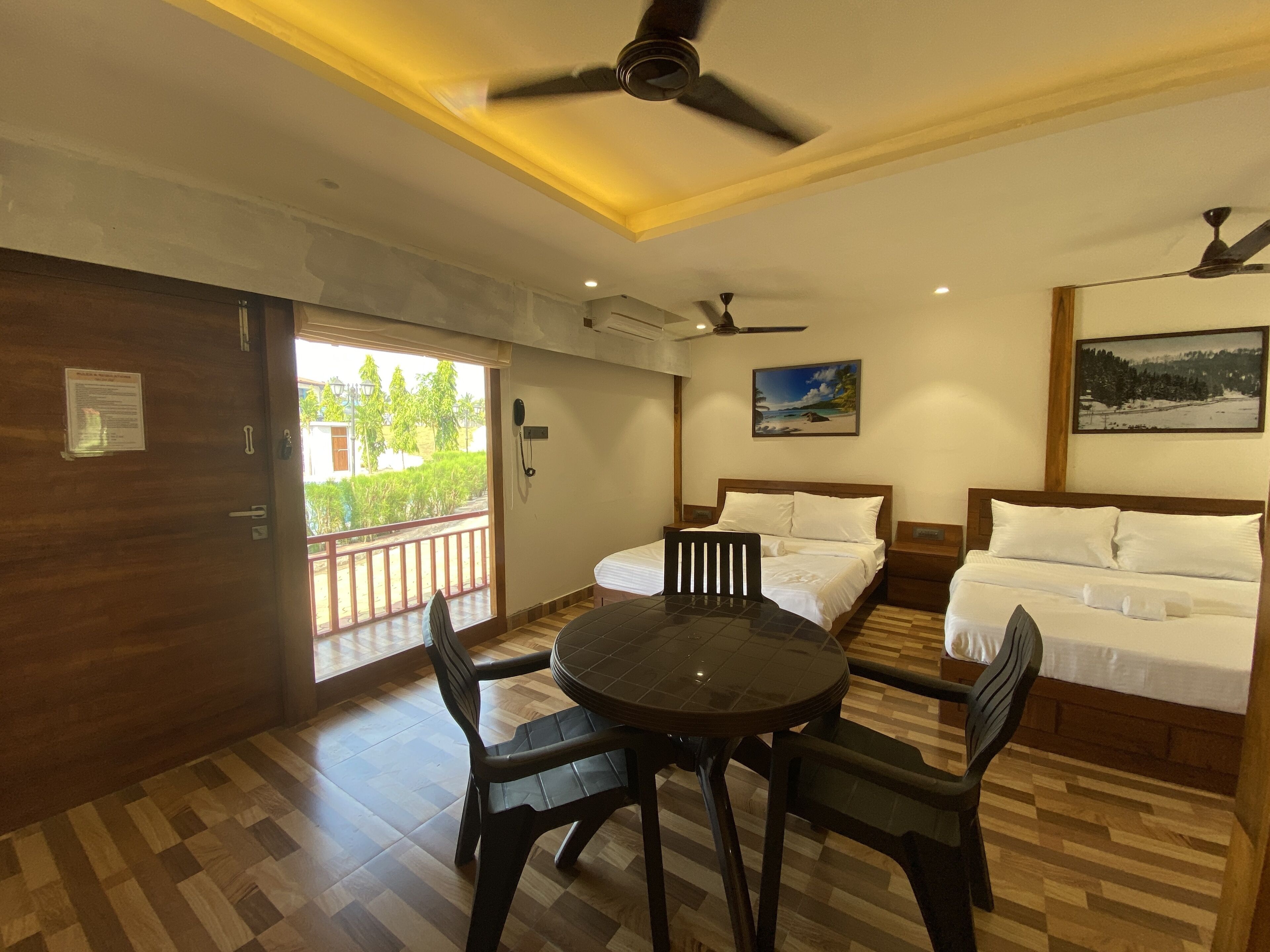 A hotel room with a balcony, two beds, a dining table with chairs, and ceiling fans.