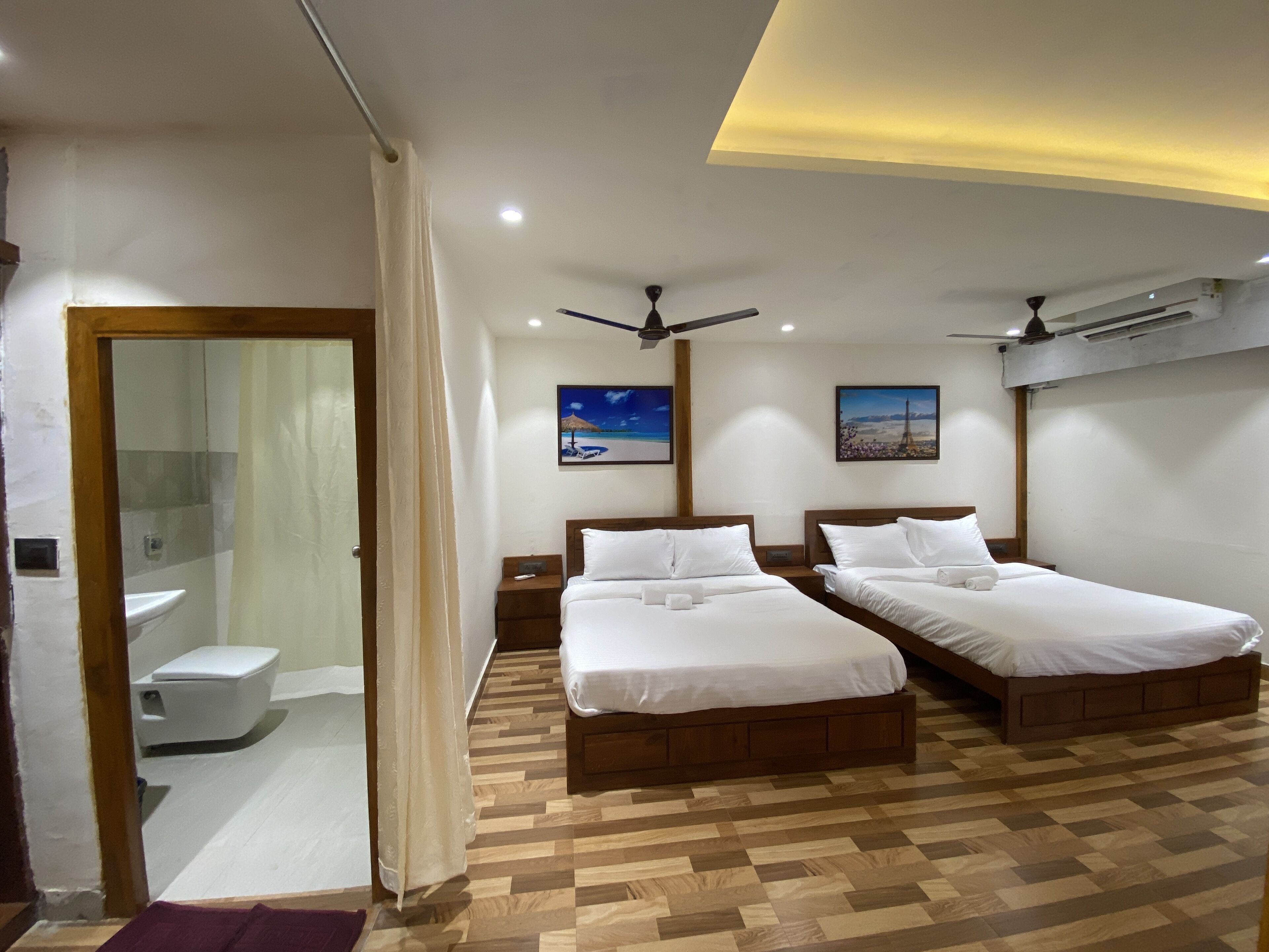 A hotel room with two beds, a wooden floor, a ceiling fan, and a painting of a cityscape.