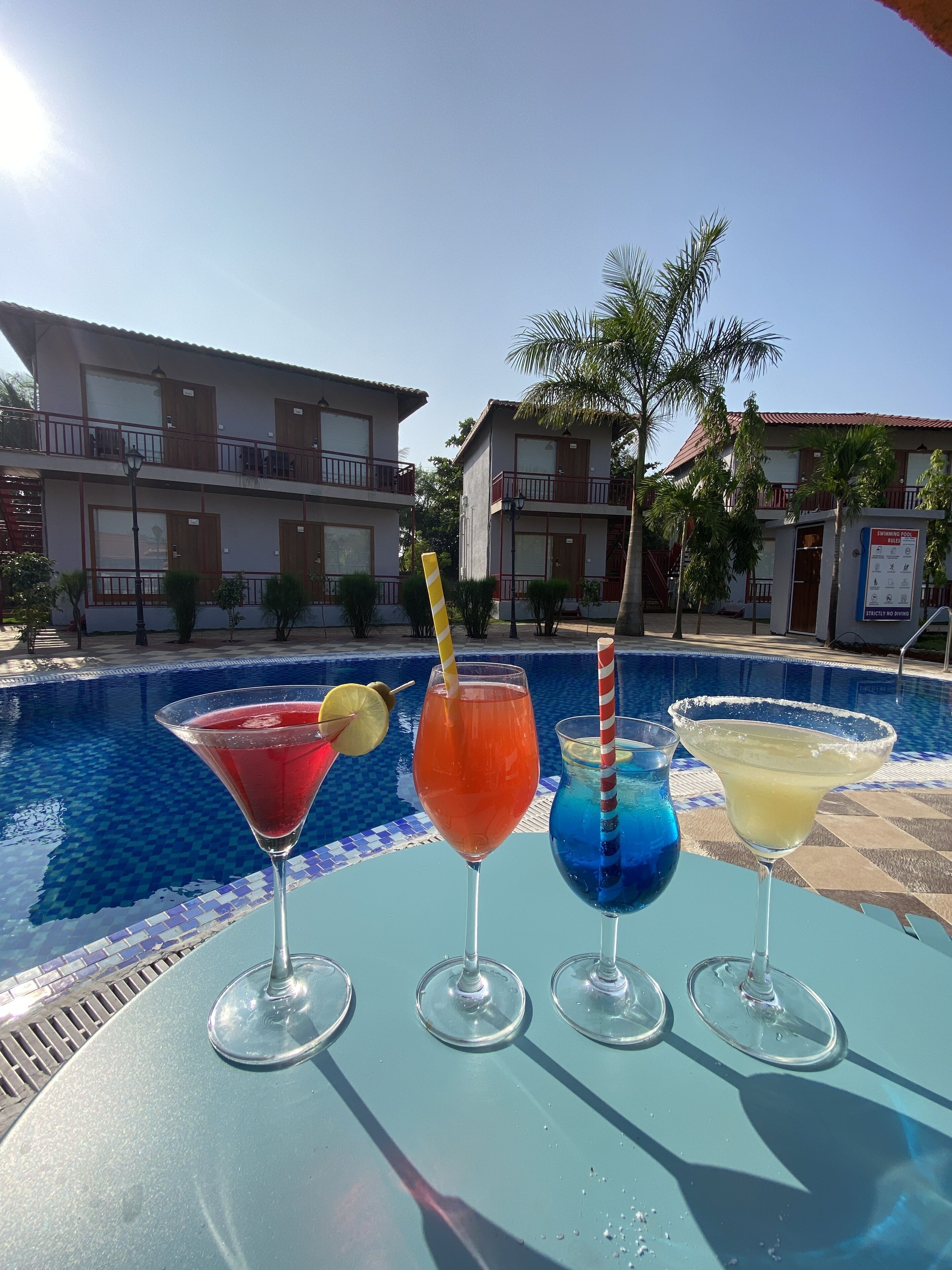 A poolside table with four drinks, a swimming pool, and a building with balconies in the background.