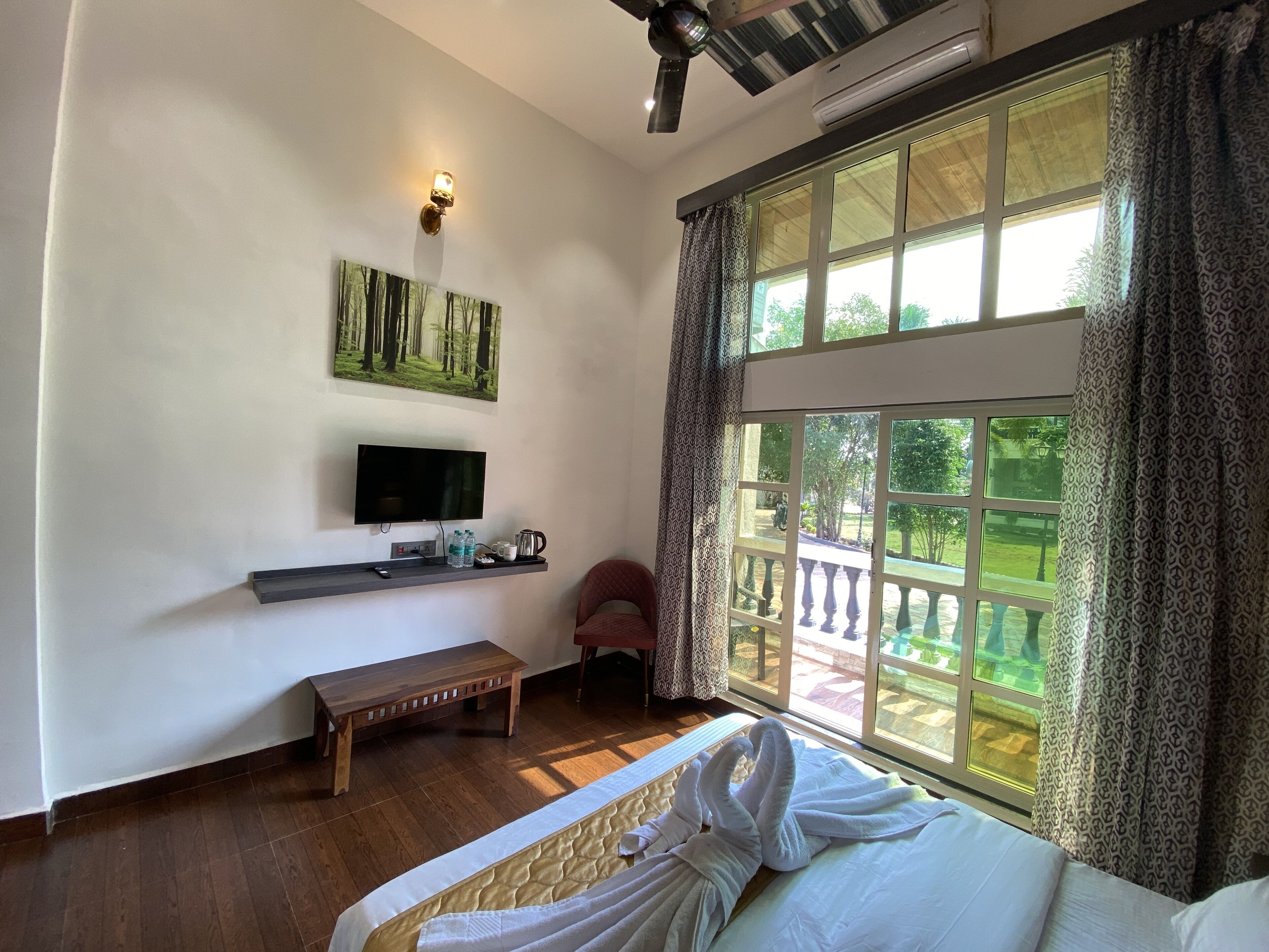 Quarto com cama, TV, um banco, janela com vista para vegetação e ventilador de teto.