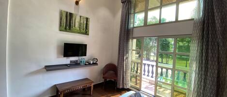 Quarto com cama, TV, um banco, janela com vista para vegetação e ventilador de teto.