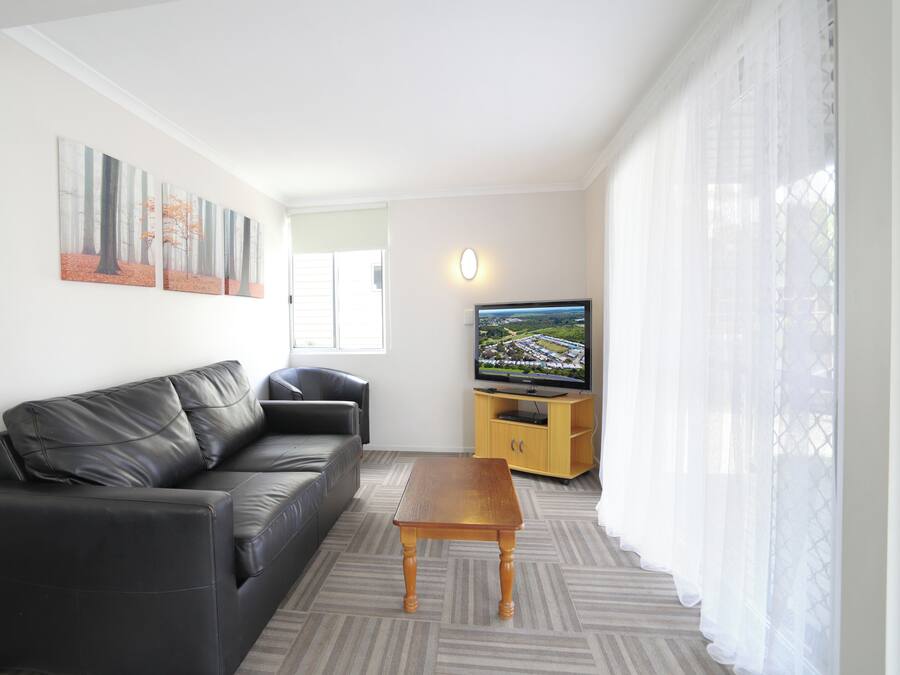 Una sala de estar moderna con un sofá de cuero negro, una mesa de centro de madera, un televisor de pantalla plana sobre un mueble de madera y un ventanal con cortinas translúcidas.