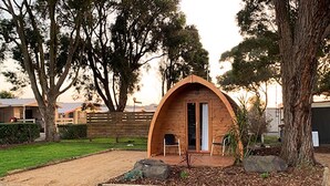 A small, single-story wooden cabin with a curved roof, a door, and a small porch with two chairs. - Wonthaggi Park Lane Holiday Park (Wonthaggi)