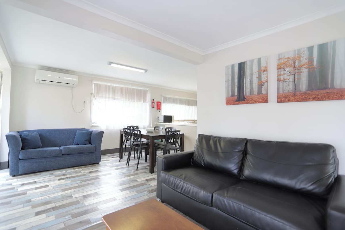 Una sala de estar con un sofá azul, un sofá de cuero negro y una mesa de centro de madera. Hay un comedor con una mesa y sillas.
