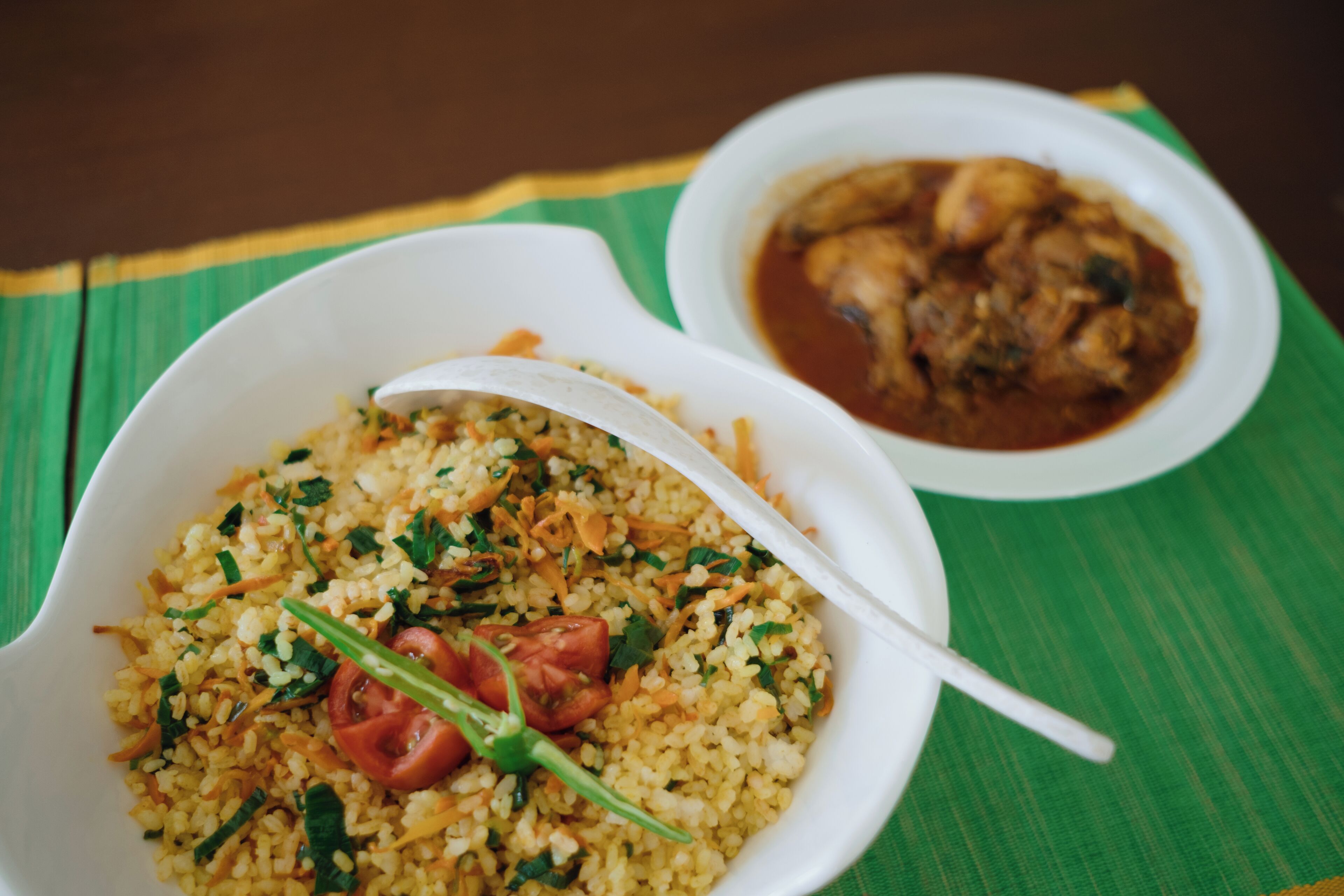A bowl of rice with vegetables and a spoon, and a bowl of stew on a green mat.