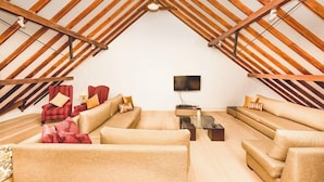 A cozy attic room with a sloped ceiling, furnished with beige sofas and a red armchair, a flat-screen TV, and a small table.