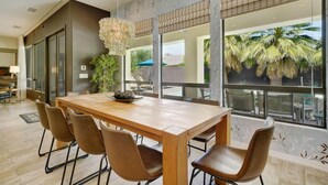 A dining area with a wooden table and brown chairs, large windows with a view of palm trees, and a hanging light fixture. - Summer Early Late Access Private Pool Spa (Indio)