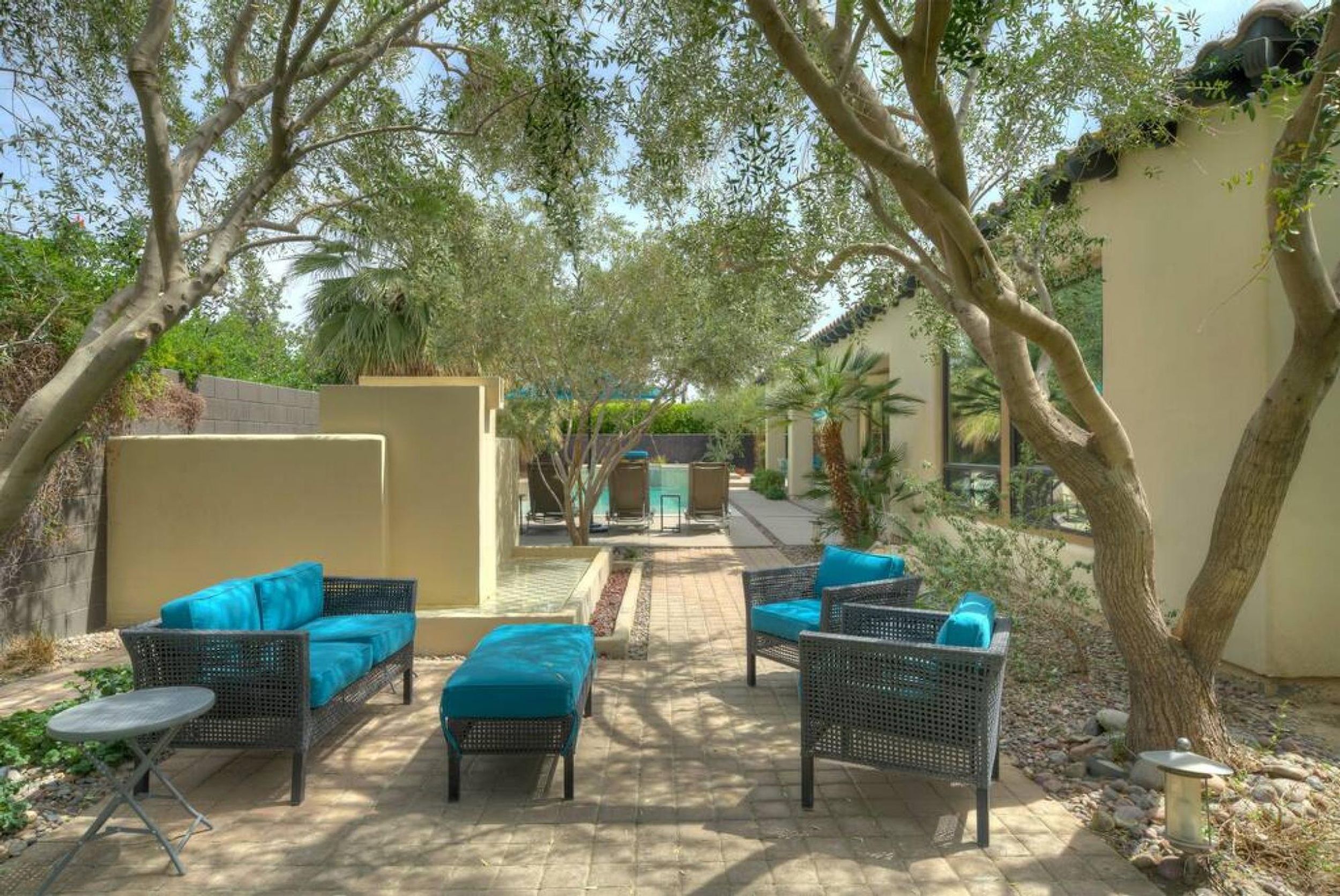An outdoor patio area with wicker furniture and blue cushions, surrounded by trees and a beige wall.