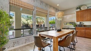 A modern kitchen with a wooden dining table, four chairs, and large windows with views of palm trees. - Summer Early Late Access Private Pool Spa (Indio)