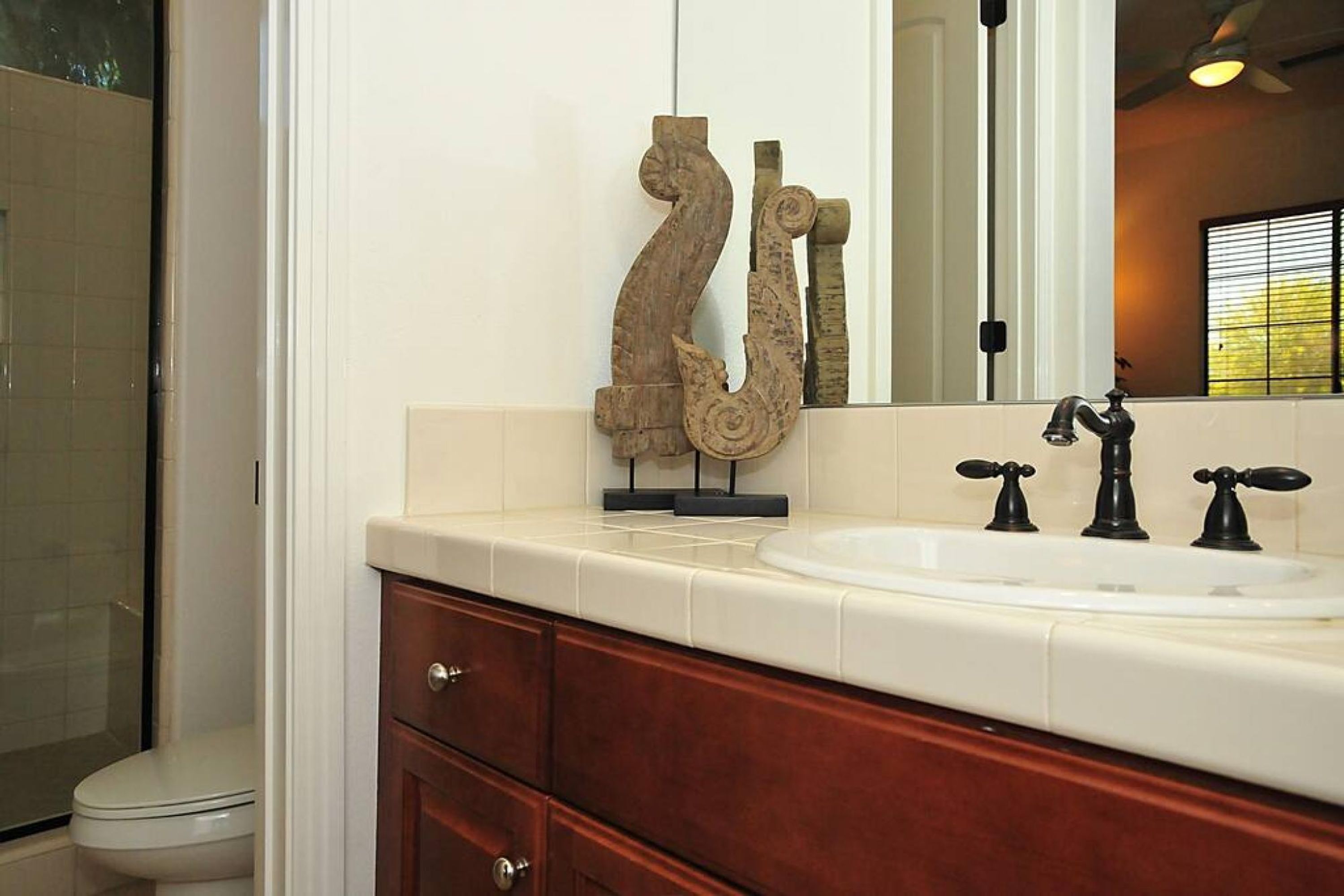 A bathroom with a white sink, wooden vanity, and a mirror reflecting a window with blinds.