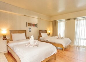 A hotel room with two beds, each with a wooden headboard, white linens, and a bedside lamp. There is a painting on the wall and a window with curtains.