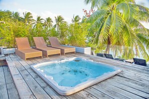 A wooden deck with a hot tub and lounge chairs, surrounded by palm trees and greenery.