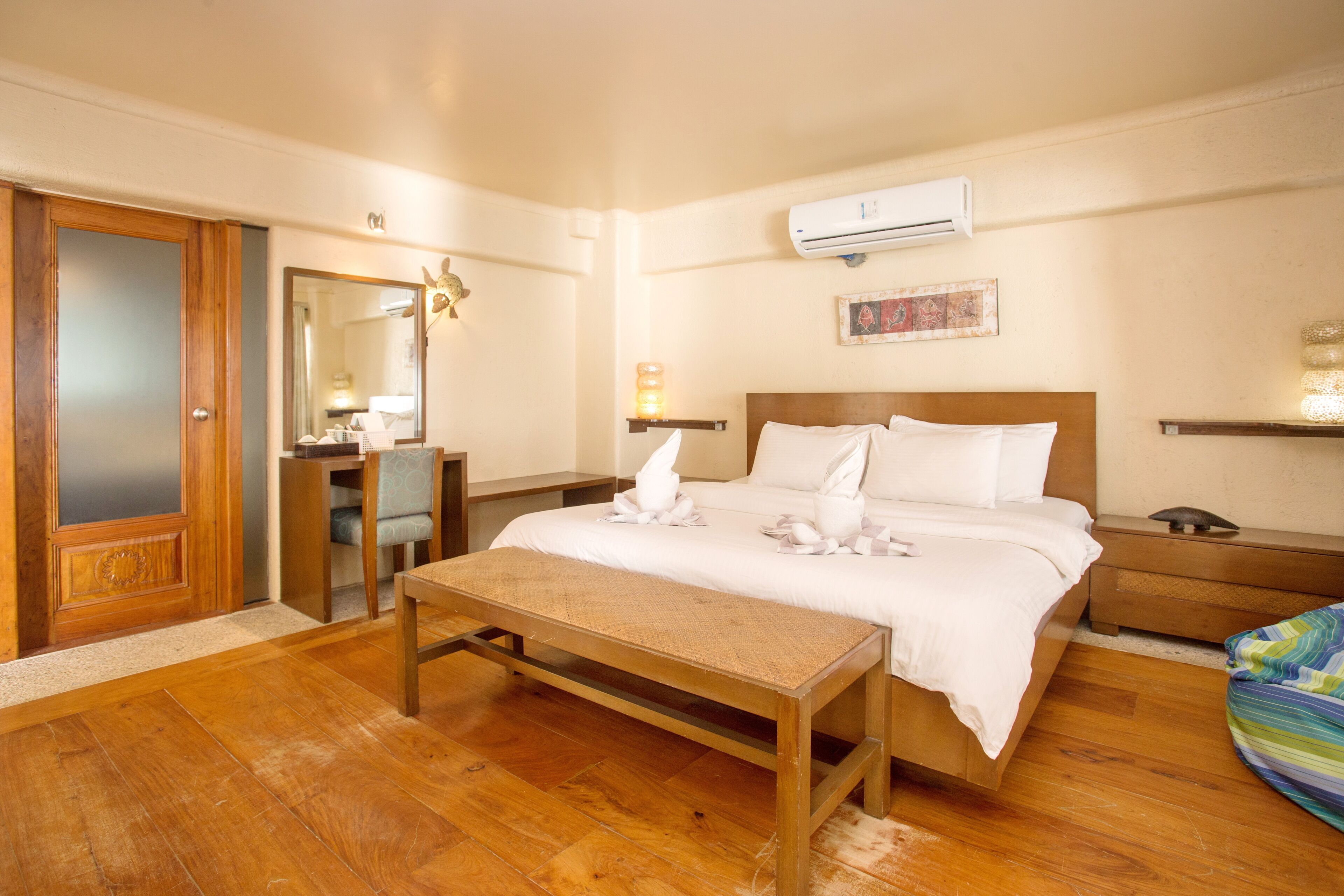 Une chambre d’hôtel avec un plancher en bois, un lit avec du linge de lit blanc, une table de chevet, une commode et un appareil de climatisation fixé au mur.