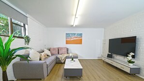 A modern living room with a grey sofa, a coffee table, a TV on a white brick wall, and a large window with a view of greenery.