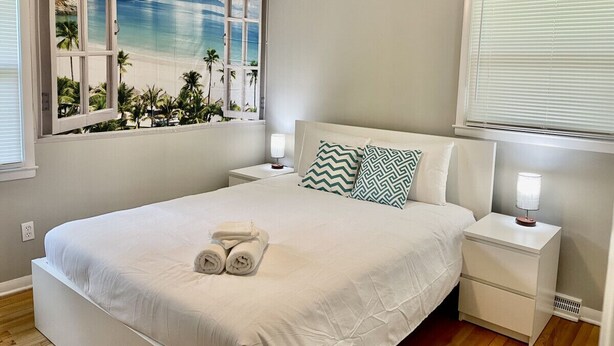 A neatly arranged bedroom with a bed, two bedside tables, and a window with a beach mural.