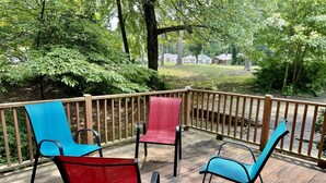 A wooden deck with four chairs, one red and three blue, overlooking a grassy area with trees and houses in the background.