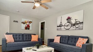 A living room with two grey sofas, a white coffee table, and a ceiling fan. There are wall decorations including a cow painting and a bicycle painting.