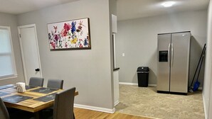 A dining area with a wooden table and chairs, a painting on the wall, a refrigerator, and a trash can.