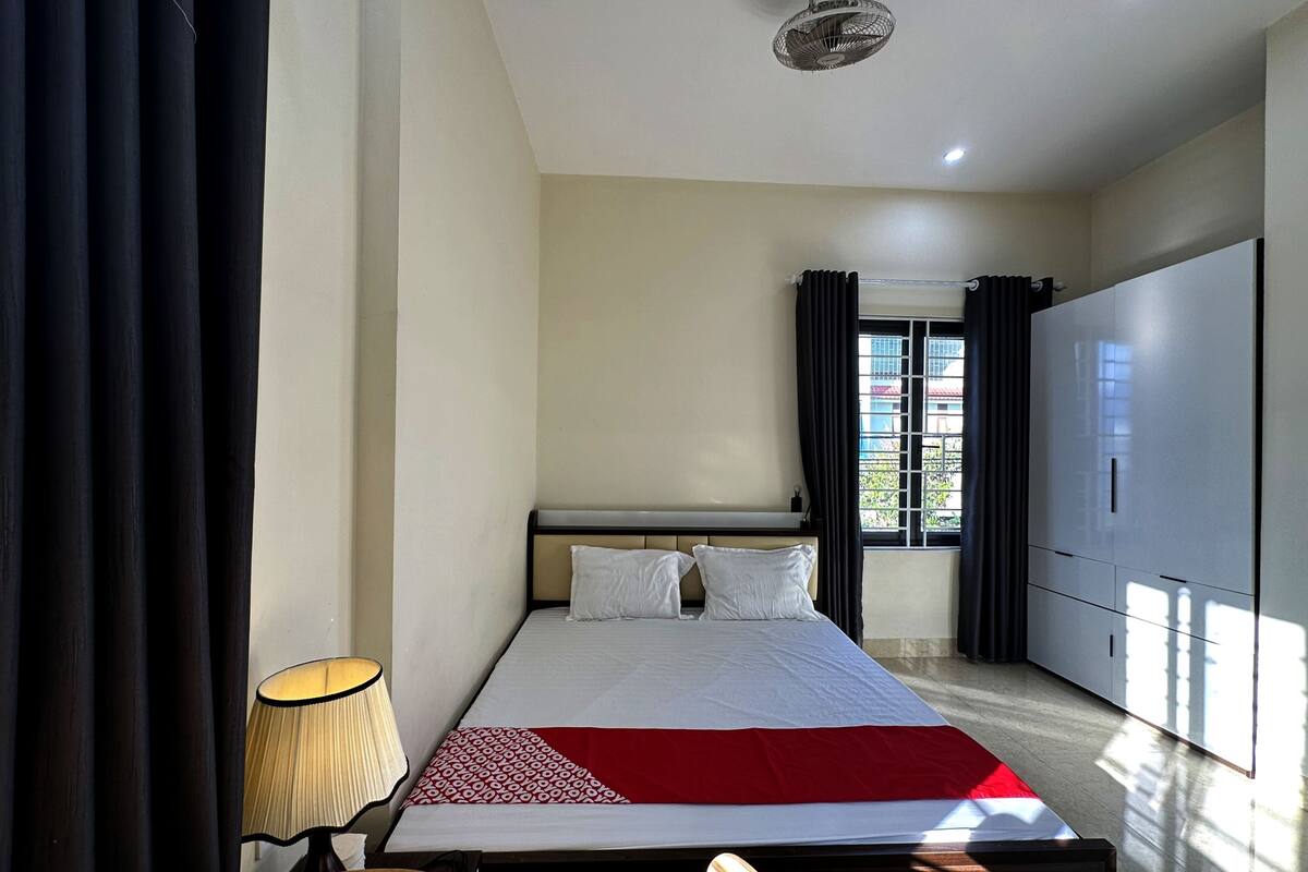 Una habitación de hotel con cama, lámpara de noche, ventana con cortinas y un armario.
