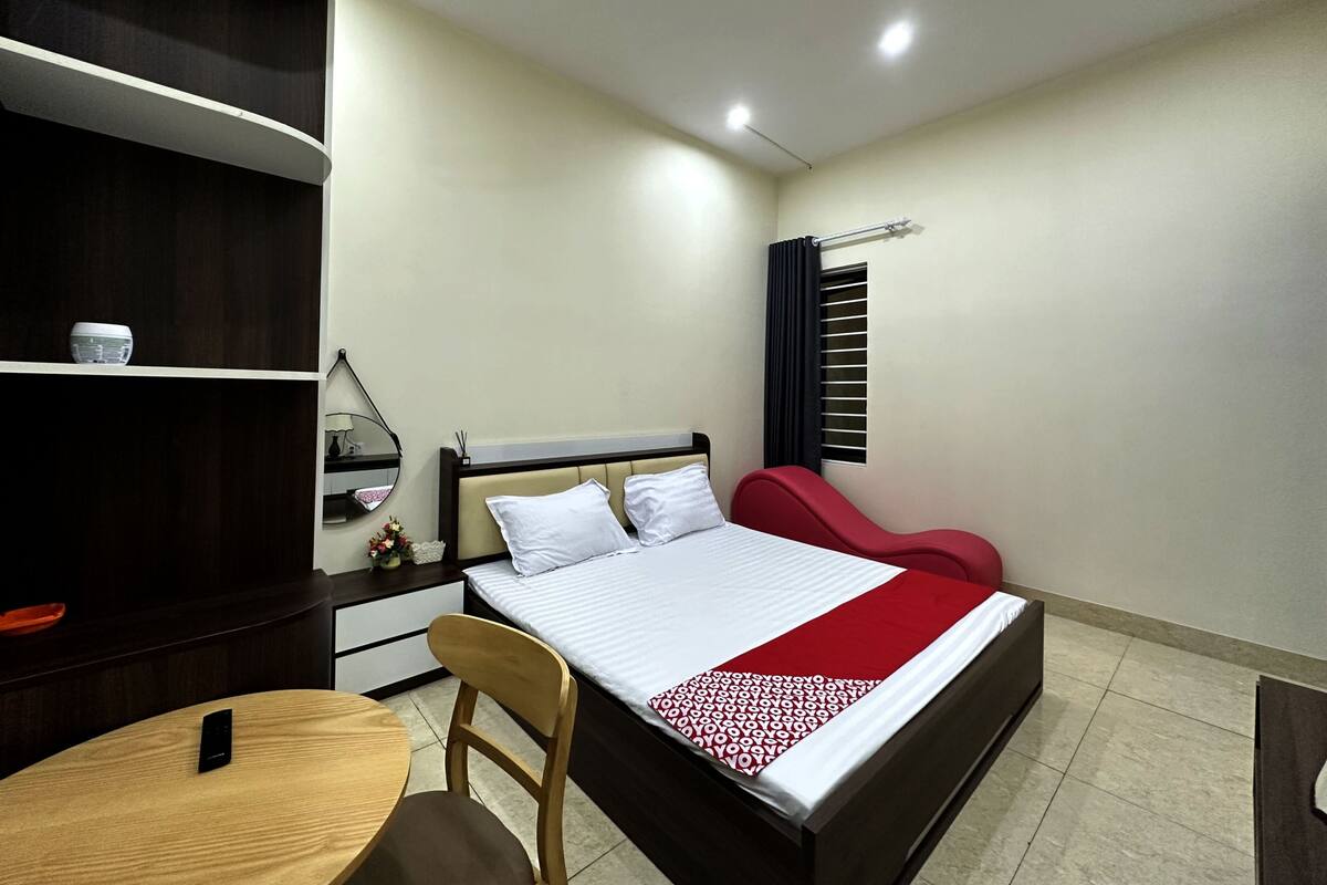 Una habitación de hotel con cama, escritorio, silla, mesita, espejo, estante y ventana con cortina.