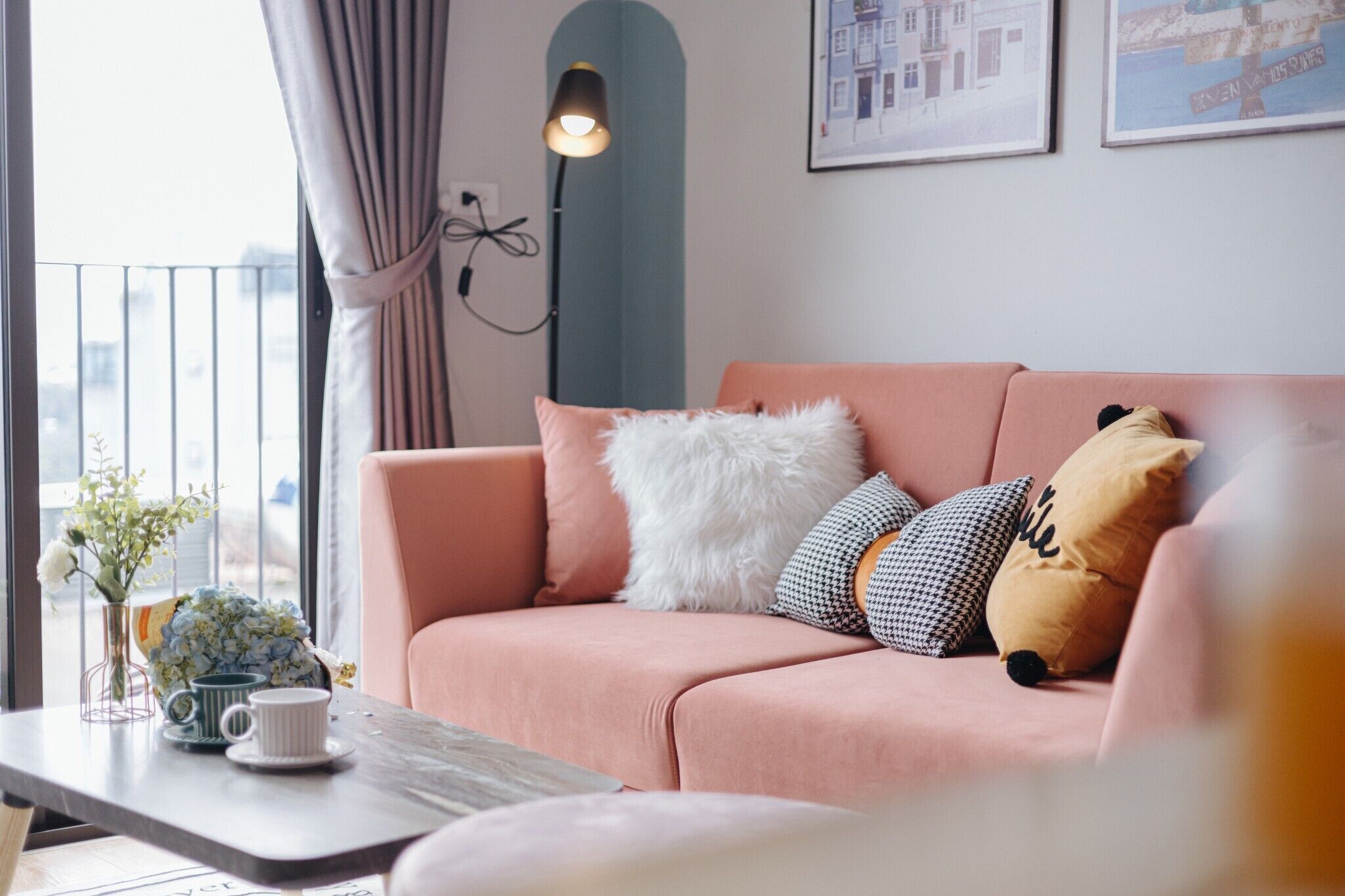 A pink sofa with various pillows, a vase with flowers, and a table with a mug and saucer.