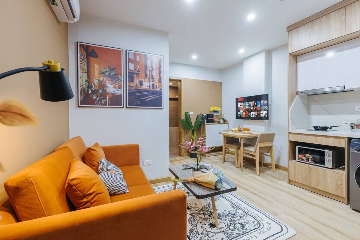 A modern living room with a sofa, a coffee table, and a dining area with a table and chairs. The kitchen area includes cabinets and a microwave.