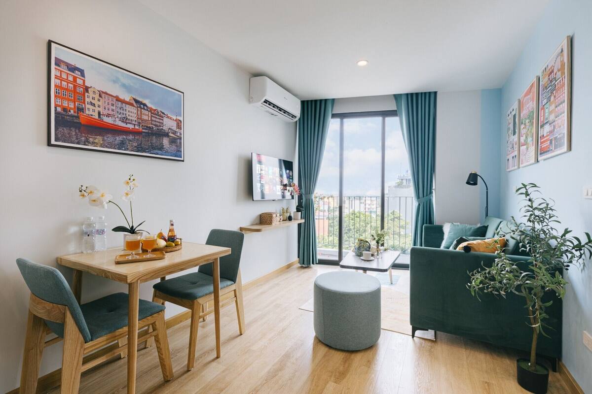 A modern living room with a dining area, a green sofa, a grey ottoman, and a balcony with a view of buildings.