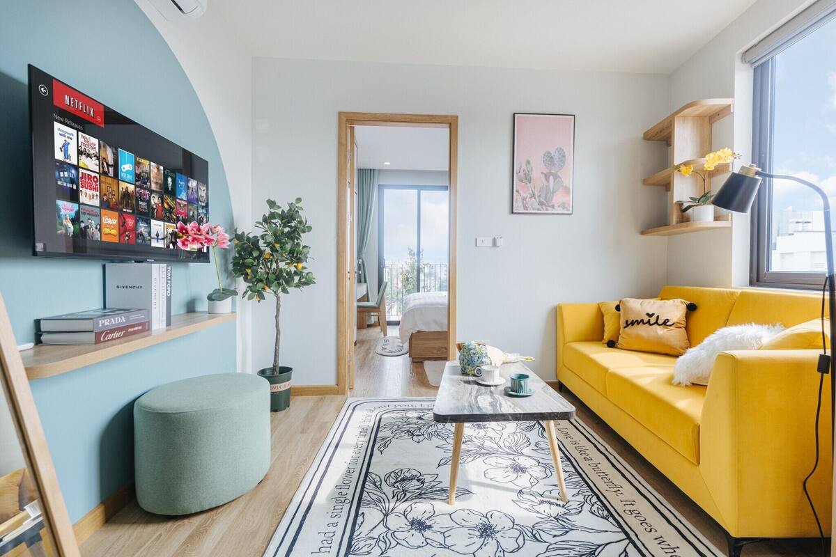 A modern living room with a yellow sofa, a flat-screen TV displaying a Netflix interface, a wooden bookshelf, and a potted plant.