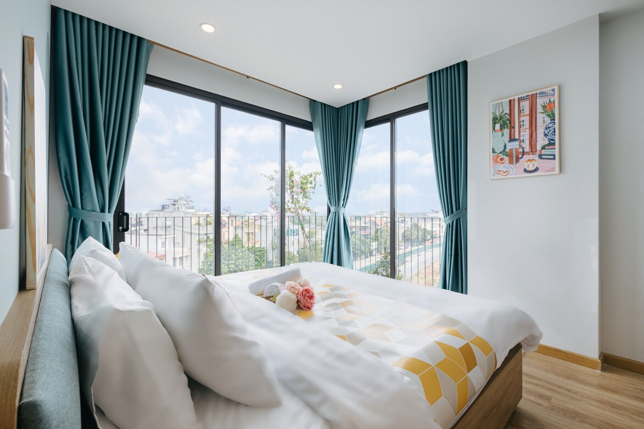A hotel room with a large bed, teal curtains, a city view, and a painting on the wall.