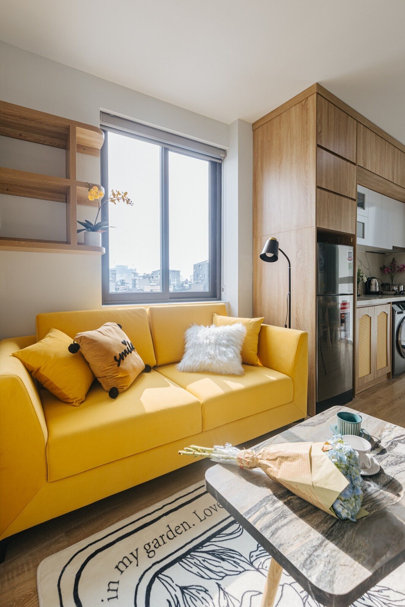 Ein modernes Wohnzimmer mit einem gelben Sofa, einem Couchtisch mit Blumenarrangements und Blick auf die Stadt durch ein großes Fenster.