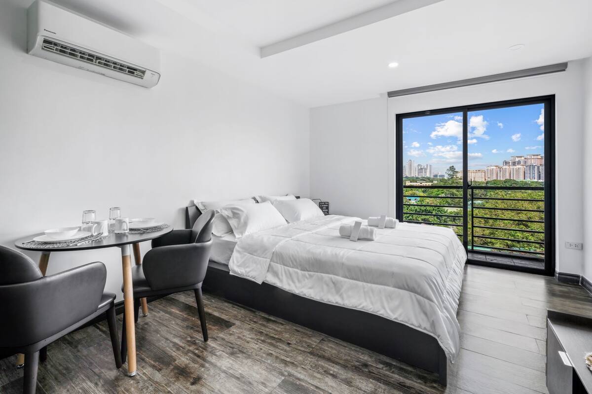 A modern hotel room with a large bed, a dining table with chairs, and a balcony with a city view.