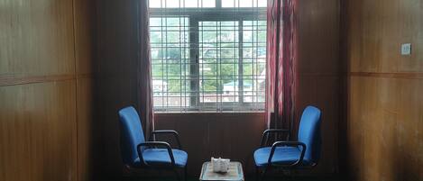 A room with two blue chairs, a small table with a book, and a window with metal bars and red curtains.