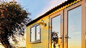 Une maison mobile jaune, dotée d’une grande fenêtre et d’une porte, est située sur un chemin de gravier, avec des marches menant à l’entrée.