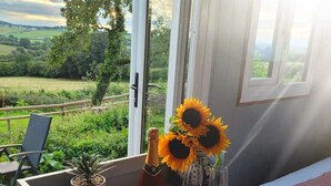 Une table à manger dressée avec des assiettes, une bouteille de champagne et un bouquet de tournesols près d’une fenêtre donnant sur un paysage verdoyant.