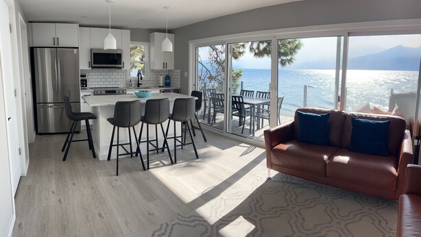 A modern kitchen with a central island, a dining area with a large table, and a sofa facing a balcony with a view of the sea.