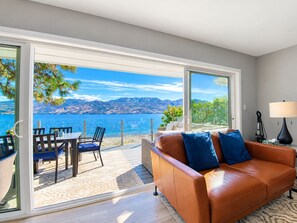 A modern living room with a large sliding glass door leading to a balcony with a view of a lake and mountains.